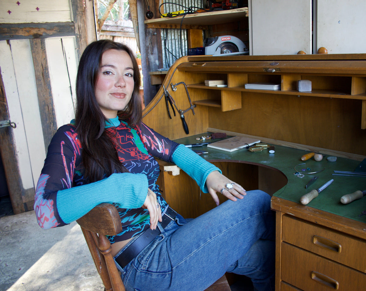 A woman with long dark hair sits in a wooden chair in front of a large wooden writing desk with tools on a green work surface. She is wearing a colorful long-sleeve top and blue jeans.
Feline Jewellery owner. Feline Jewellery 
