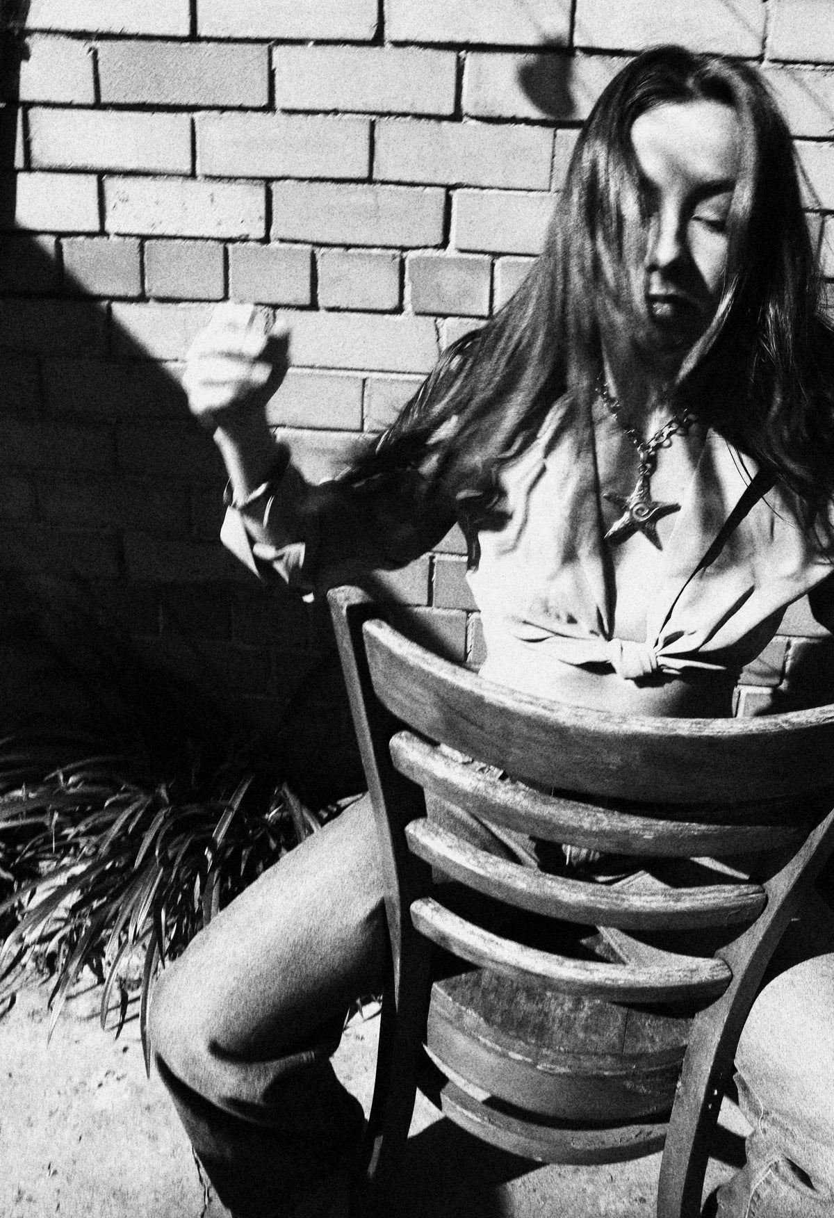 Black and white photograph of a woman seated on a wooden chair against a brick wall, wearing a light-colored top and dark pants, with strong shadows across the scene.
Feline Jewellery owner Feline Jewellery